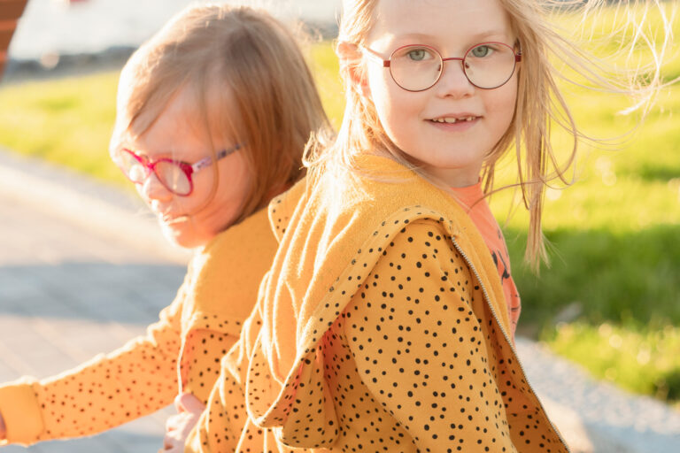 Kaksi silmälasipäistä tyttöä oransseissa huppareissa istuvat sataman penkillä kesäiltana.