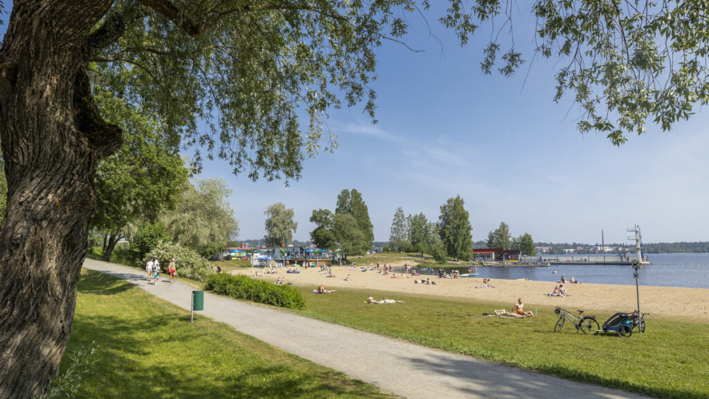 Kesäinen päivä Väinölänniemen uimarannalla. Etualalla hiekkatie, jonka takana nurmialue ja uimaranta.