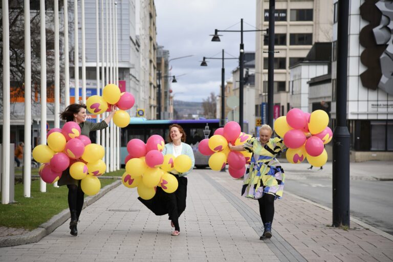 Kolme henkilö värillisten ilmapallojen kanssa kadulla.