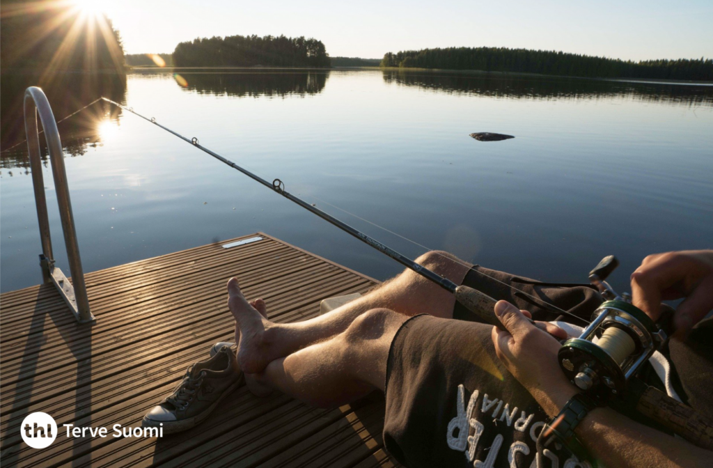 Henkilö istuu laiturilla onkimassa kauniissa kesäillassa. Järvi on täysin tyyni.