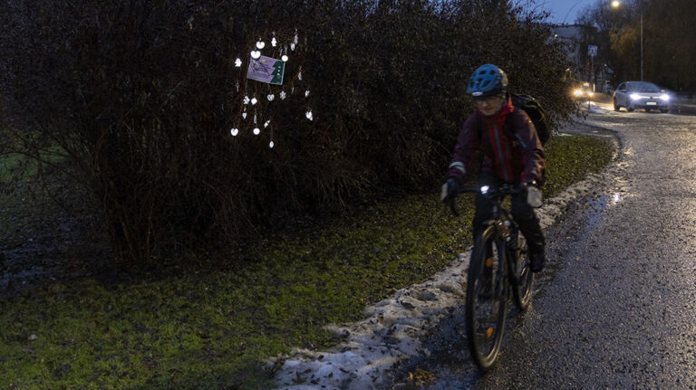 Pyöräilijä ajaa pyöräilyväylän reunassa aamuhämärässä. Taustalla pensas, jossa roikkuu oksilla heijastimia.