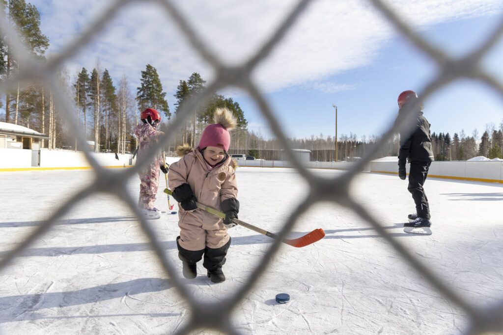 Pieni tyttö on kaukalon jäällä jääkiekkomailan kanssa, kuva otettu suojaverkon läpi
