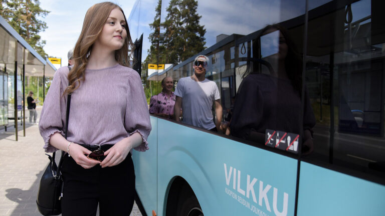 Kuopion seudun joukkoliikenteen linja-auto oikealla ja vasemmalla nuori naishenkilö kävelemässä.
