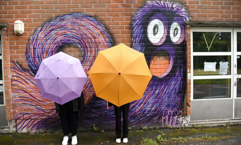 Kuvassa tiiliseinään on maalattu spraylla violetti hauska karvaisen näköinen olento. Kaksi henkilöä seisoo sen edessä sateenvarjot kasvojensa ja ylävartaloiden peitteenä.