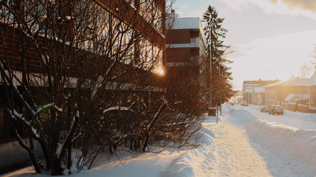 Auringon valo kajastaa oikeasta laidasta lumiselle kadulle, jonka vasemmassa reunassa ruskea kerrostalo.