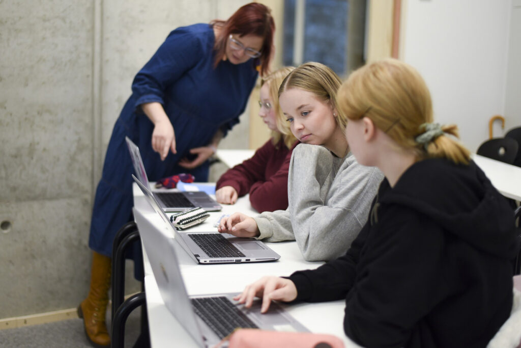Naishenkilö kumarassa osoittaa tietokoneen näyttöä ja opastaa. Kolme nuorta naista istuu pöydän ääressä tietokoneella luokassa.