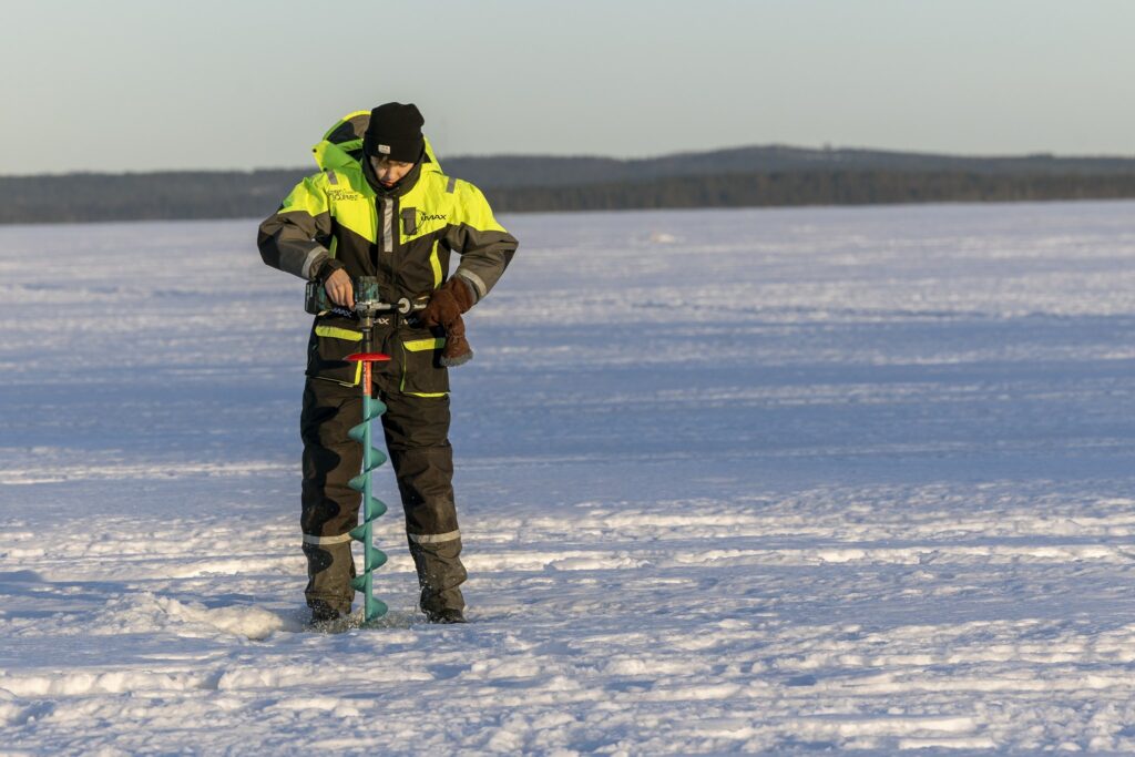 Mieshenkilö kairaa pilkkireikää jäällä. Taustalla, kaukana näkyy metsää.