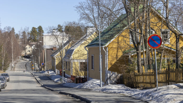 Keltaisia taloja jalankulkuväylän oikealla puolella. Keväinen aurinkoinen sää, sulat tiet, joiden reunoilla hieman lunta.