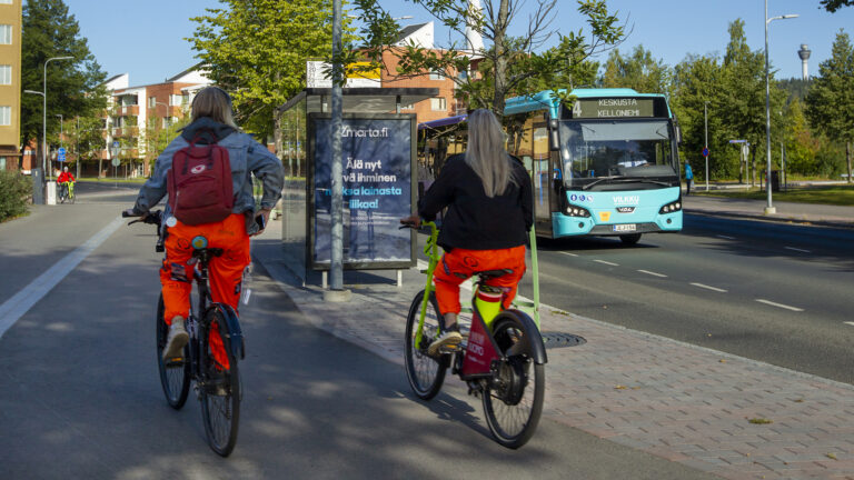 Etualalla kaksi henkilöä pyöräilee Vilkku-fillareilla, edessään bussipysäkki ja oikeassa laidassa linja-auto.