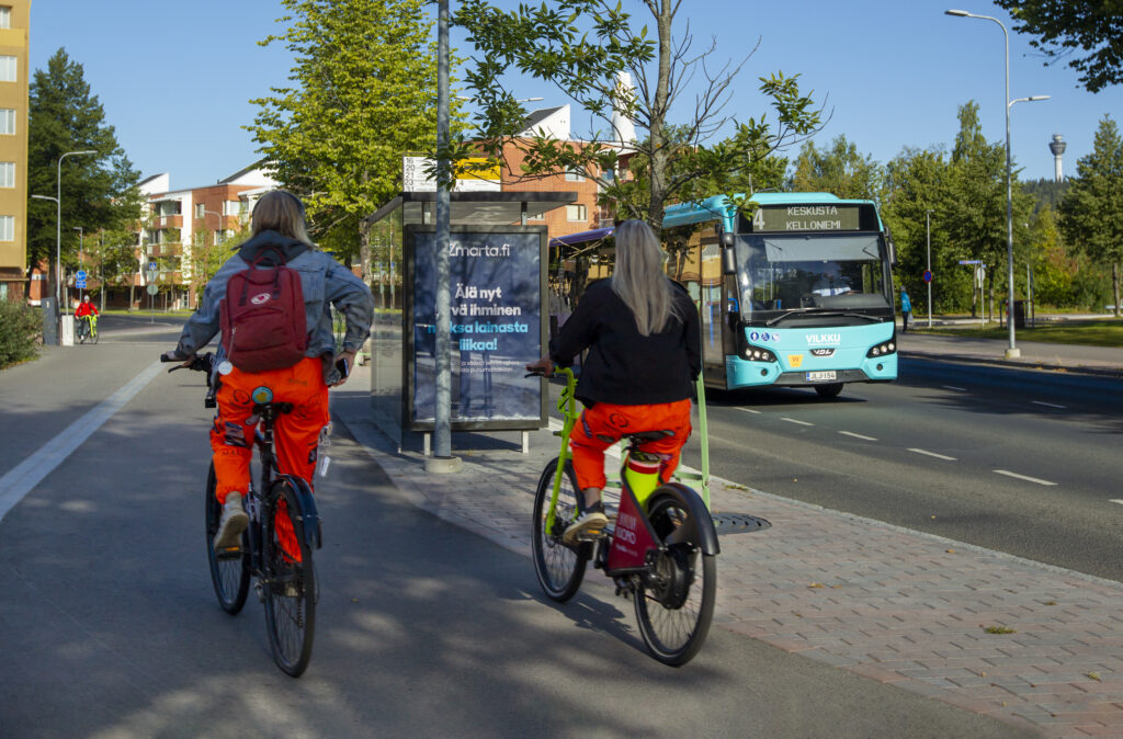 Kesäisellä kadulla pyöräilee kaksi henkilöä, selkä kuvaajaan päin, sähköpyörillä. Oikealla puolella vastaan tulee Vilkku-bussi. Kohteiden välissä näkyy bussipysäkki.