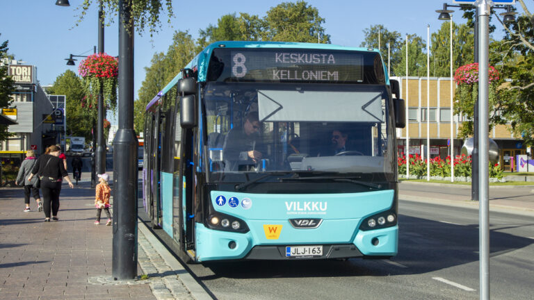 Turkoosin värinen linja-auto pysähtyneenä torin laidalle kesäisenä aurinkoisena päivänä.