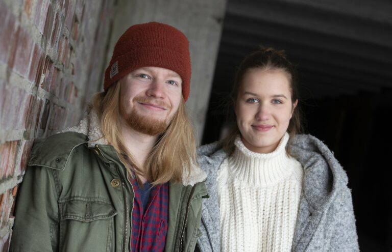Nuori mies ja nainen katsovat kohti kameraa ja hymyilevät hieman. Taustalla tiiliseinää ja betonipylväs. Nuorella miehellä päällä vihreä takki ja punainen pipo. Nuorella naisella päällä valkoinen villapaita ja harmaa takki.