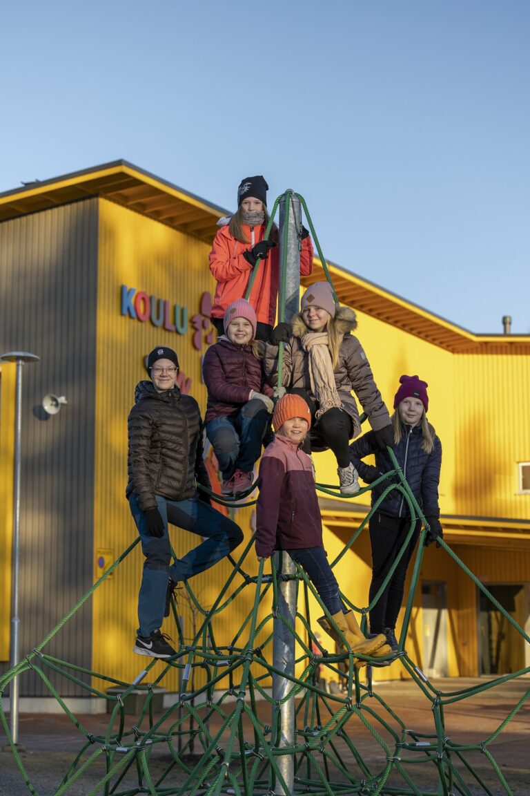 Kuusi lasta on kiivennyt kiipeilypyramidiin ja katsovat kohti kameraa iloisina. Osa lapsista istuu ja osa seisoo köysillä.