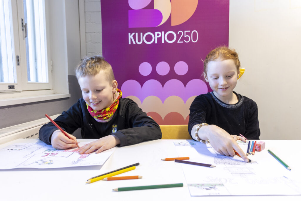 Poika ja tyttö piirtävät pöydän äärellä. Takana näkyy Kuopio 250-juhlavuoden mainos.