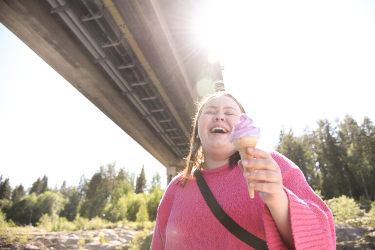 Nauravainen ihminen jäätelö kädessä sillan alla, aurinko luo valopisaroita hänen ylleen