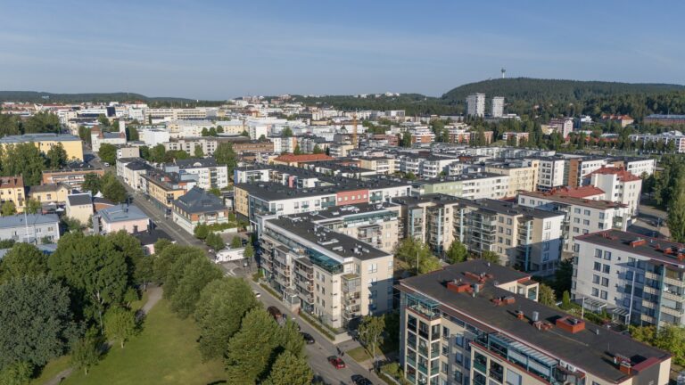 Sininen taivas taustalla, kerrostaloja sekä vihreää kasvillisuutta etualalla.